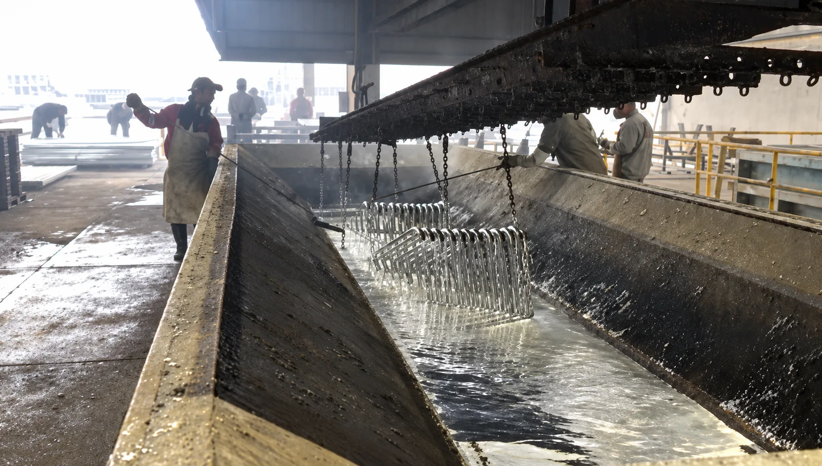 Grades de isolamento presas a correntes, mergulhadas num tanque comprido da galvanização, com operários dos dois lados